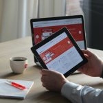 Business professional using a tablet and laptop with a hot drink, focusing on digital content review.