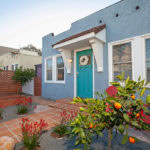 Exterior of a blue bungalow at 3764 Alabama in North Park with a teal front door, terracotta tile path, red Adirondack chairs, and citrus trees in a landscaped front yard.