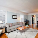 Sun-drenched living room at 3764 Alabama in North Park with a grey sofa, hardwood floors, a geometric rug, and large windows with white blinds.