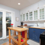 Modern kitchen at 3764 Alabama St in North Park featuring a butcher block island, navy blue lower cabinets, and white shaker upper cabinets with glass fronts, situated next to sliding glass doors that lead to a private deck.
