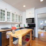 Bright, modern kitchen at 3764 Alabama St in North Park featuring white shaker upper cabinets with glass fronts, butcher block countertops, navy blue lower cabinetry, and original hardwood floors.
