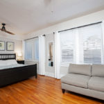 Bright primary bedroom at 3764 Alabama St in North Park featuring original hardwood floors, a black wooden bed frame, and a comfortable grey sofa near large windows.