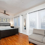 Large primary bedroom at 3764 Alabama St in North Park featuring a black wood bed frame, original hardwood floors, a neutral love seat, and bright windows with white blinds and curtains.