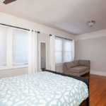 Spacious primary bedroom at 3764 Alabama St in North Park featuring warm hardwood floors, large windows with white blinds and curtains, and a cozy seating area with a neutral sofa.