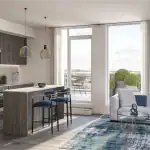 Modern open-concept kitchen and living area at The Wilmot, showcasing wood-grain cabinetry, a large island with blue velvet stools, and bright floor-to-ceiling windows leading to a private balcony.