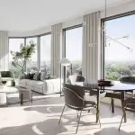 An open-concept luxury suite interior at The Greenwich showing a bright living and dining area with panoramic floor-to-ceiling windows, modern minimalist furniture, and views of a lush green landscape.