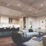 Open-concept great room at King’s Calling showing a modern kitchen with light wood cabinetry, a marble backsplash, and a living area with a coffered ceiling, fireplace, and contemporary furniture.