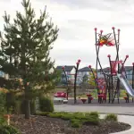 A modern playground in Cy Becker park featuring a unique red and black climbing structure with a silver slide, surrounded by green grass, trees, and residential houses in the background.