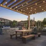Close-up of the outdoor amenity area at 77 McQueen townhomes, featuring a couple dining under a wooden pergola with string lights, a stone-base BBQ station, and a children's playground in the background.