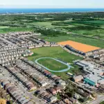 Aerial view of the On The Ridge residential community in Stoney Creek, showing rows of modern houses, a central green space with a running track and sports field, and an orange-highlighted area indicating a future development site near the lakefront.