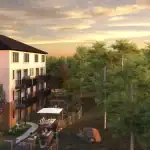 Exterior rendering of Riverbank Place townhomes at sunset, showcasing private balconies, fenced backyards with patio furniture, and lush green landscaping against a golden sky.