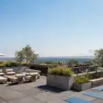 Rooftop patio amenity space at Black Birch Heights Condos featuring outdoor lounge seating, dining areas under umbrellas, concrete planters with greenery, and yoga mats looking out over a scenic view.