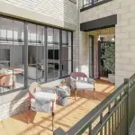 Private balcony rendering at OG Urban Towns featuring wood plank flooring, modern outdoor chairs, a small marble side table, and a large multi-pane glass window looking into the open-concept living area.