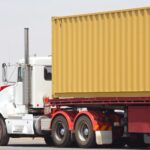 A white semi truck pulling a yellow shipping container on a flatbed trailer, representing commercial trucking business and logistics operations.