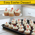 Multiple rows of cupcakes with white frosting and red jelly bean toppers arranged on a rustic table with tulips in the background.