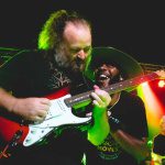 A lively live music performance featuring guitarists on stage with colorful lighting.