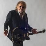 A guitar-playing man with sunglasses and curly hair, dressed in a black suit.
