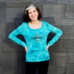 A woman smiling in a turquoise tie-dye sweatshirt standing against a textured black wall.