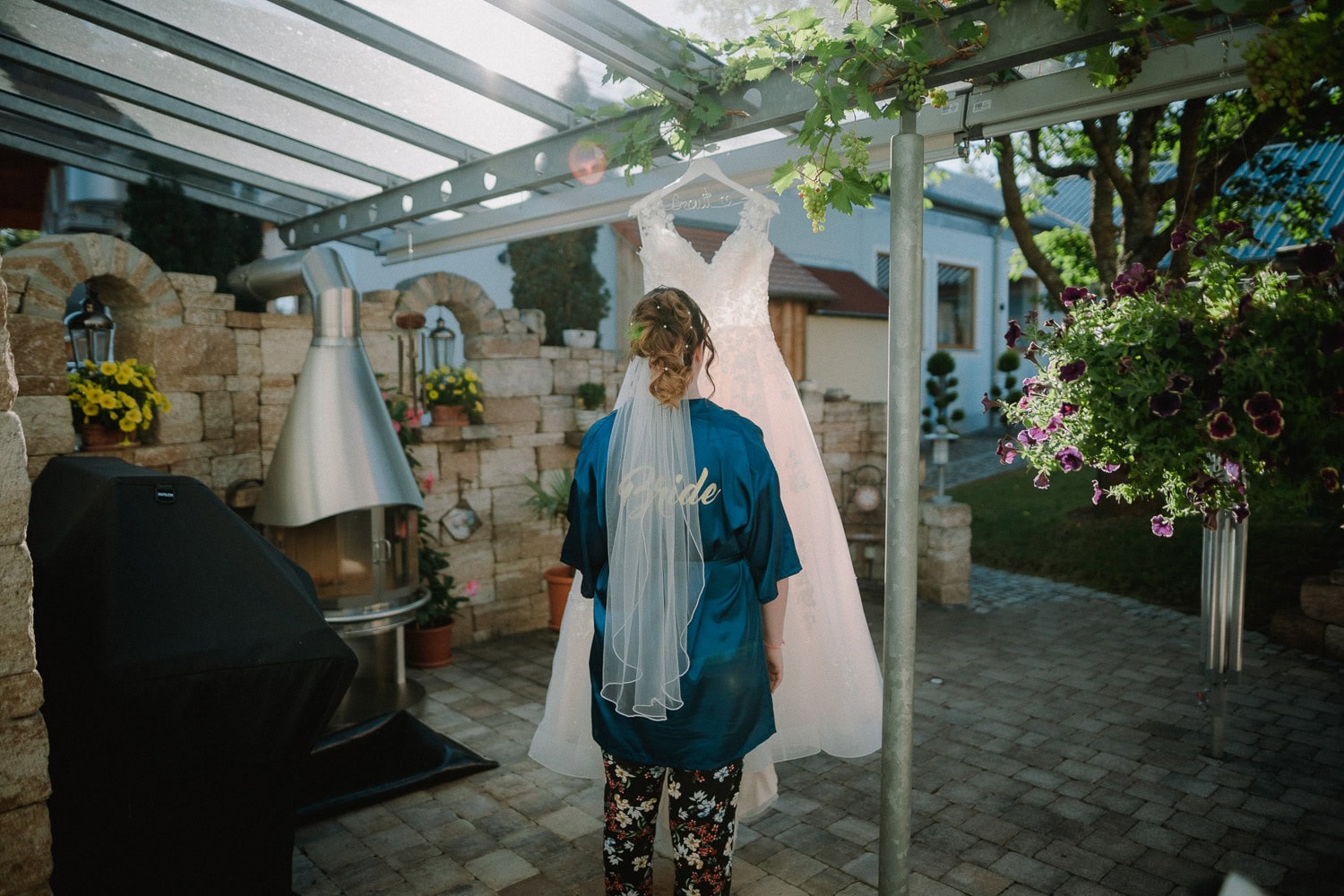 Eine Braut in einem blauen Gewand mit der Aufschrift „Braut“ auf dem Rücken steht unter einer Pergola und betrachtet ihr Hochzeitskleid, das daneben hängt. Aufgenommen von Hochzeitsfotograf Kallmünz, schaffen Steinmauern und Topfpflanzen eine heitere Atmosphäre im Freien.