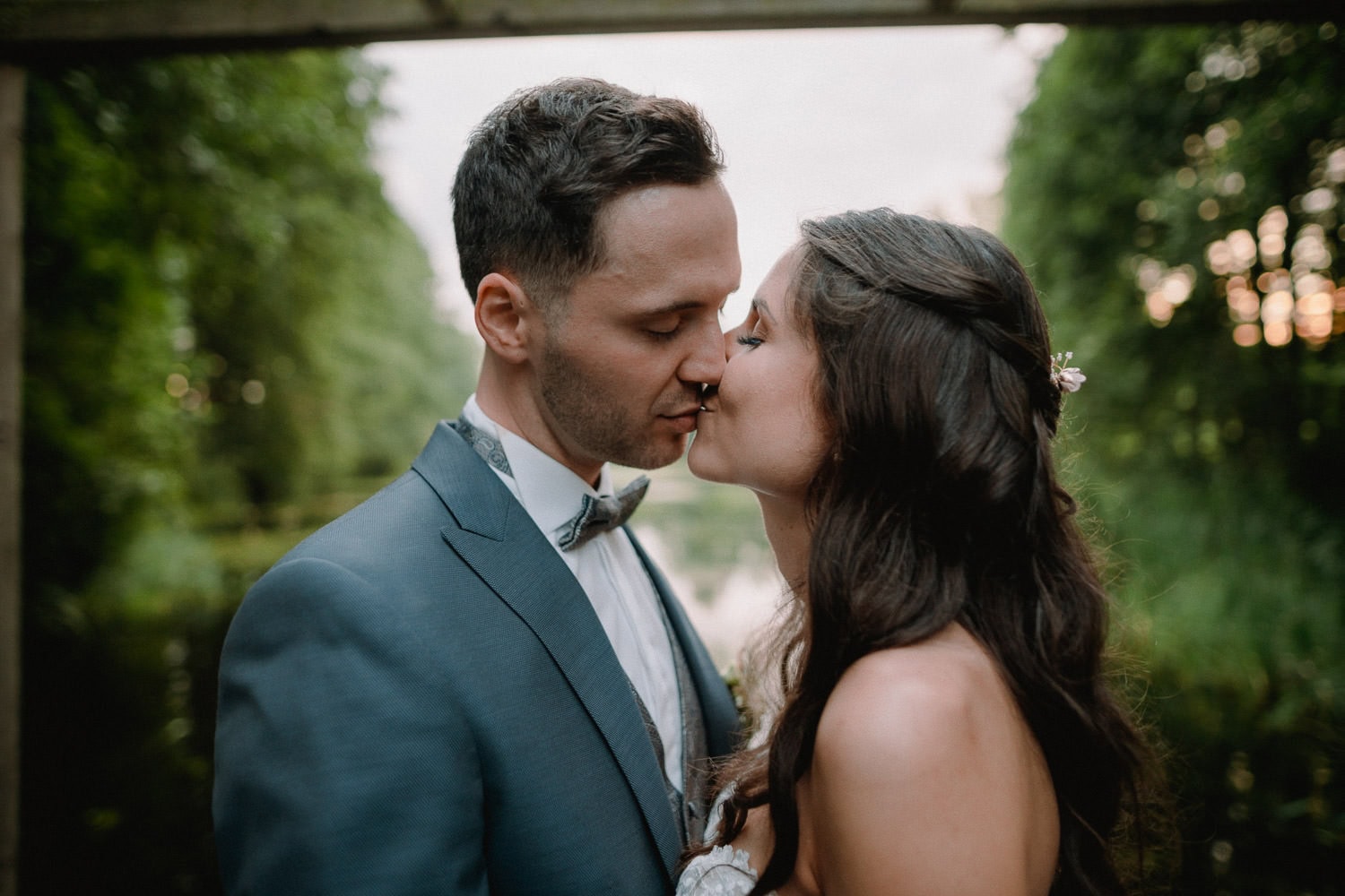 Braut und Bräutigam küssen sich im Freien auf dem Hochzeitsschloss Pirkensee, umgeben von üppigem Grün. Der Bräutigam trägt einen grauen Anzug und eine Fliege, während die Braut langes, dunkles Haar mit floralen Akzenten hat. Dahinter schaffen verschwommene Bäume und sanftes Licht eine romantische Kulisse.