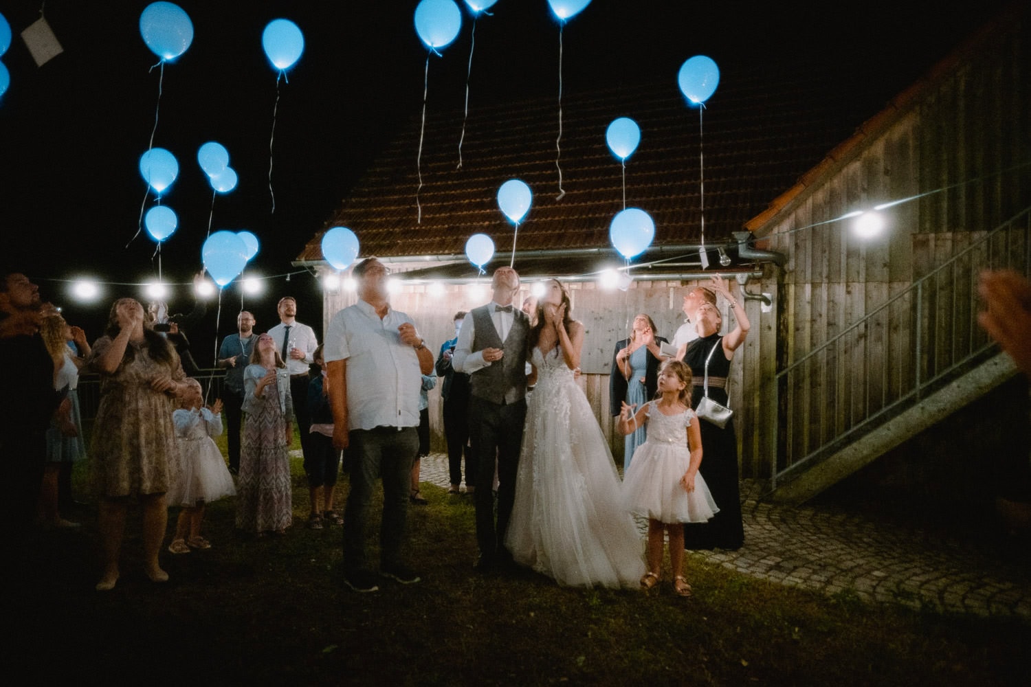 Braut und Bräutigam stehen unter dem Nachthimmel bei Hochzeit Schloss Pirkensee, umgeben von Freunden und Familie, die leuchtend blaue Luftballons halten. Die Gruppe wirkt fröhlich, mit einem rustikalen Holzgebäude im Hintergrund und Lichterketten, die die festliche Atmosphäre verstärken.
