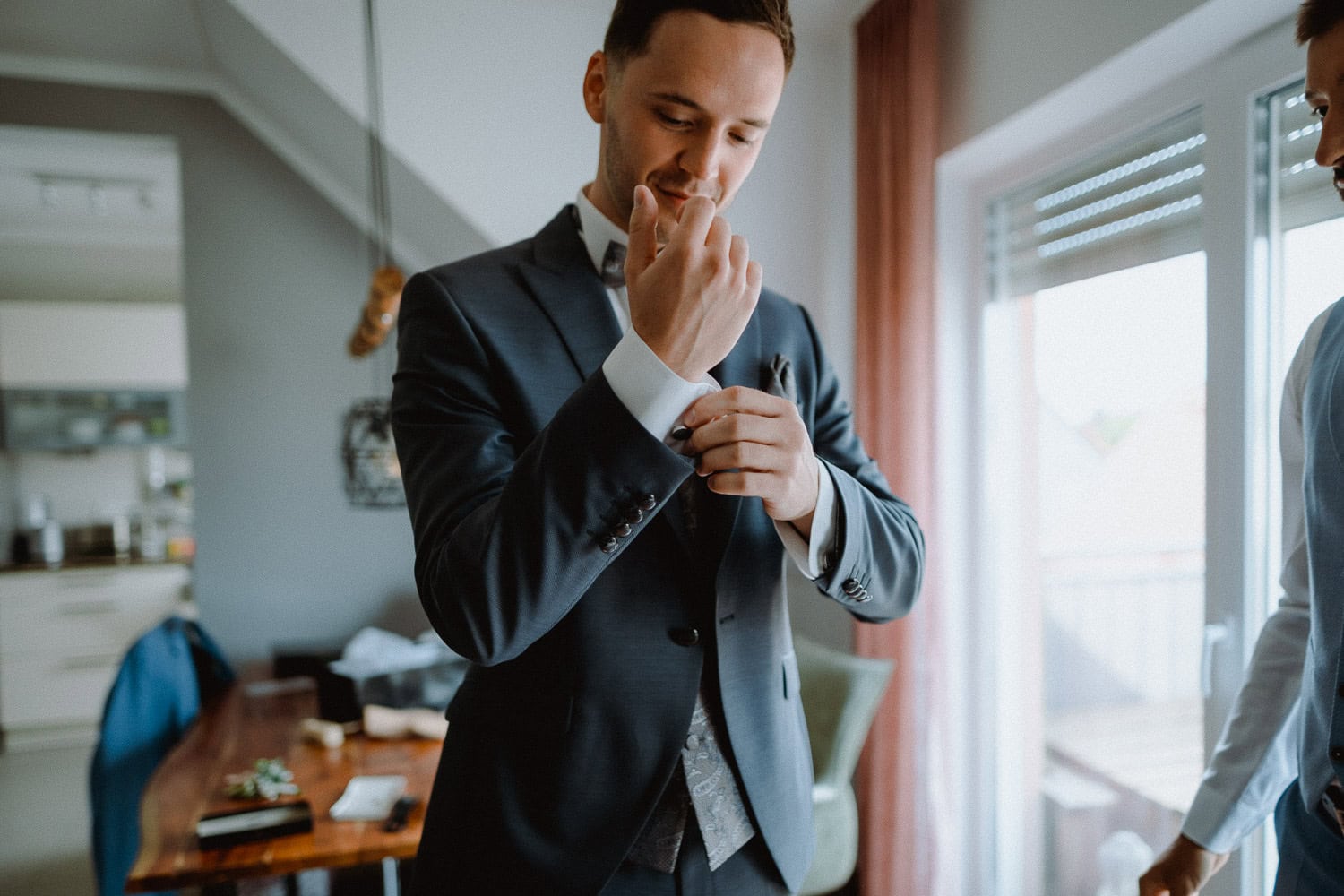 Ein Mann im Anzug rückt seine Manschettenknöpfe in einem modernen, gut beleuchteten Raum zurecht. Er steht neben einem Holztisch und einem Fenster mit halb heruntergelassenen Jalousien und bereitet sich auf die Hochzeit im Schloss Pirkensee vor. Eine weitere Person, teilweise sichtbar, steht in der Nähe. Der Raum ist gemütlich und modern eingerichtet.
