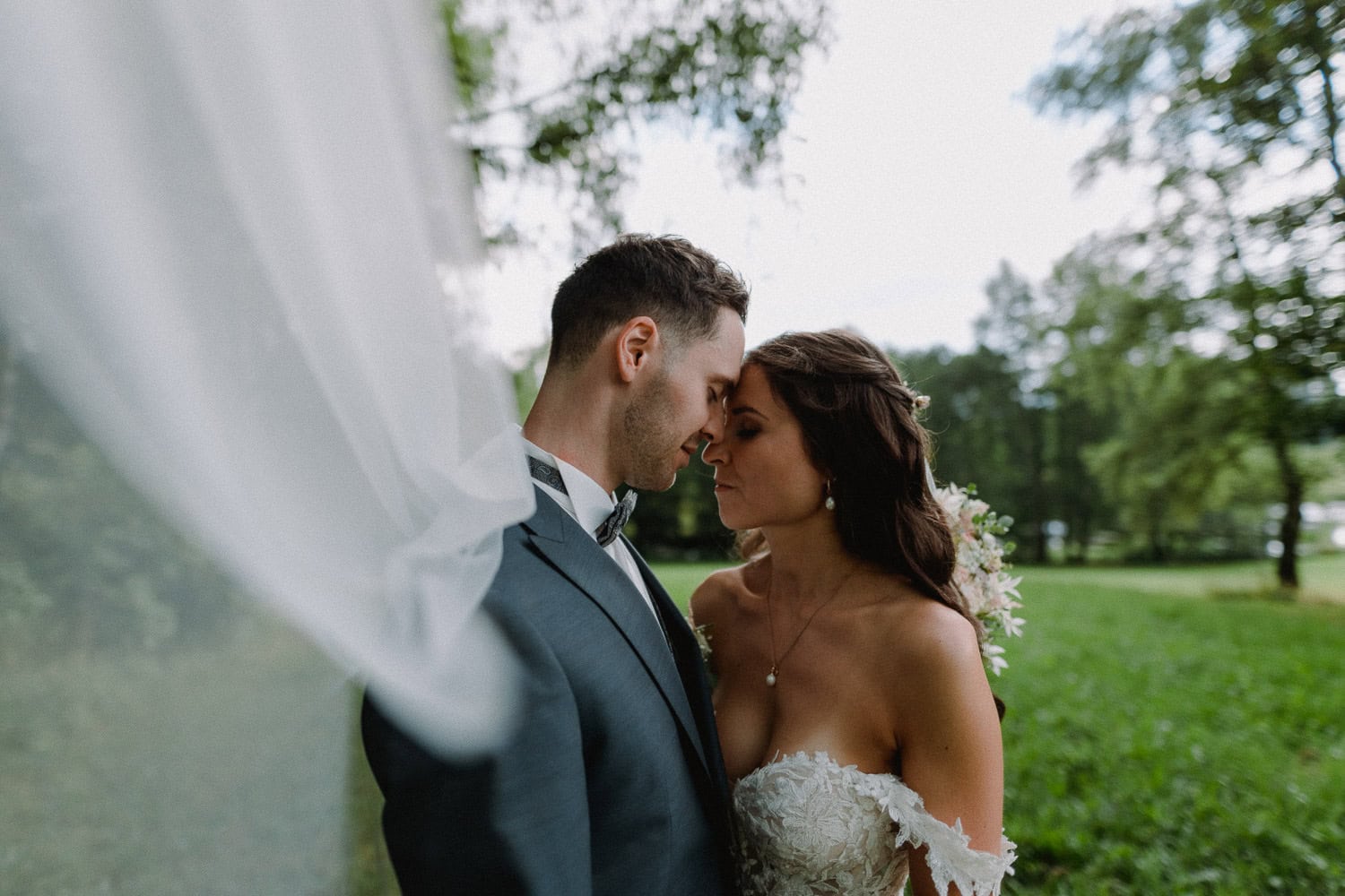 In einer üppigen, grünen Umgebung im Freien von Schloss Pirkensee steht ein Paar eng beieinander im Hochzeitskleid, die Stirnen berühren sich. Der Bräutigam trägt einen dunklen Anzug, während die Braut in einem weißen, schulterfreien Kleid und Schleier glänzt. An ihrem Hochzeitstag sind sie von der Ruhe der Natur umgeben.