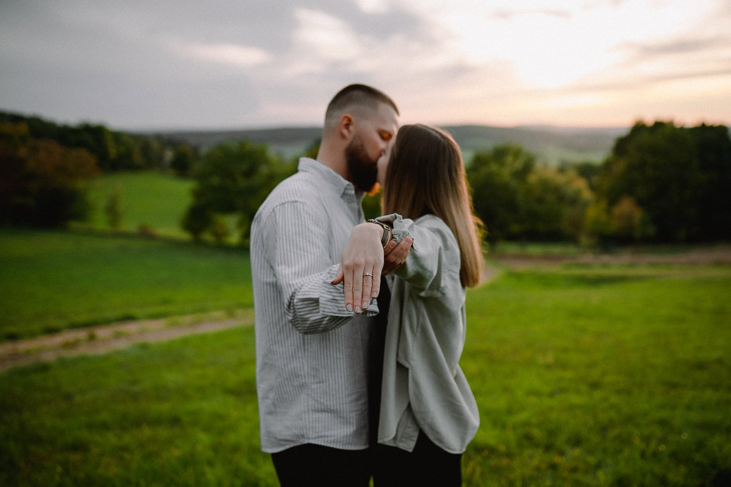Ein Paar steht bei Sonnenuntergang auf einer grünen Wiese, umarmt sich und zeigt stolz einen Ring an der Hand der Frau, was ihre Verlobung andeutet. Die Kulisse besteht aus sanften Hügeln und einem teilweise bewölkten Himmel und fängt die Essenz eines romantischen Paarshootings perfekt ein.