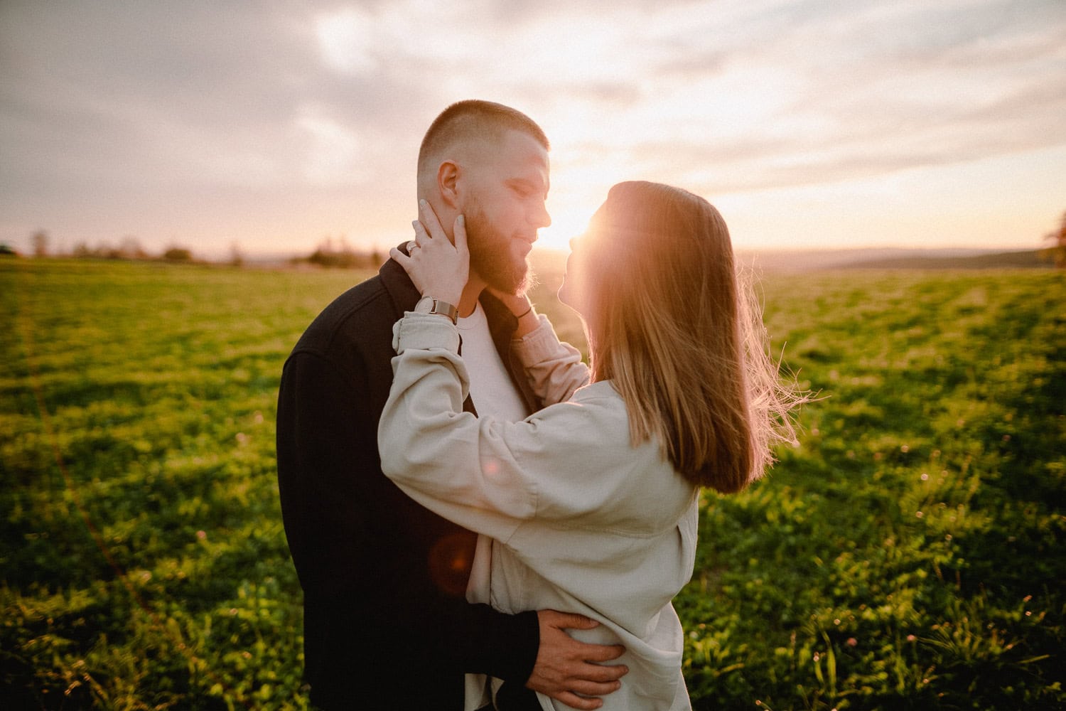 Auf einer sonnenbeschienenen Wiese erlebt ein Paar einen intimen Moment während seiner Verlobung beim Paarshooting. Die Frau berührt sanft das Gesicht des Mannes, während die Sonne hinter ihnen untergeht und einen warmen Schein über die grasbedeckte Landschaft und ihre liebevolle Umarmung wirft.
