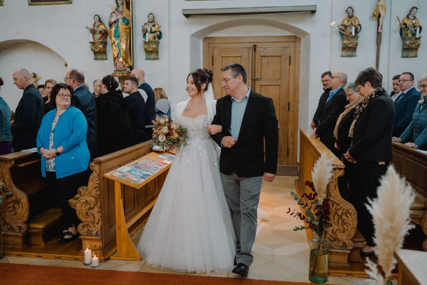 Eine Braut im weißen Brautkleid schreitet mit einem Mann im Anzug den Gang des Hammerschlosses Stodl Schmidmühlen entlang. Umgeben von Gästen in den Kirchenbänken ist die Szene warm und fröhlich, Kerzen und Blumenarrangements schmücken den Raum wunderschön.