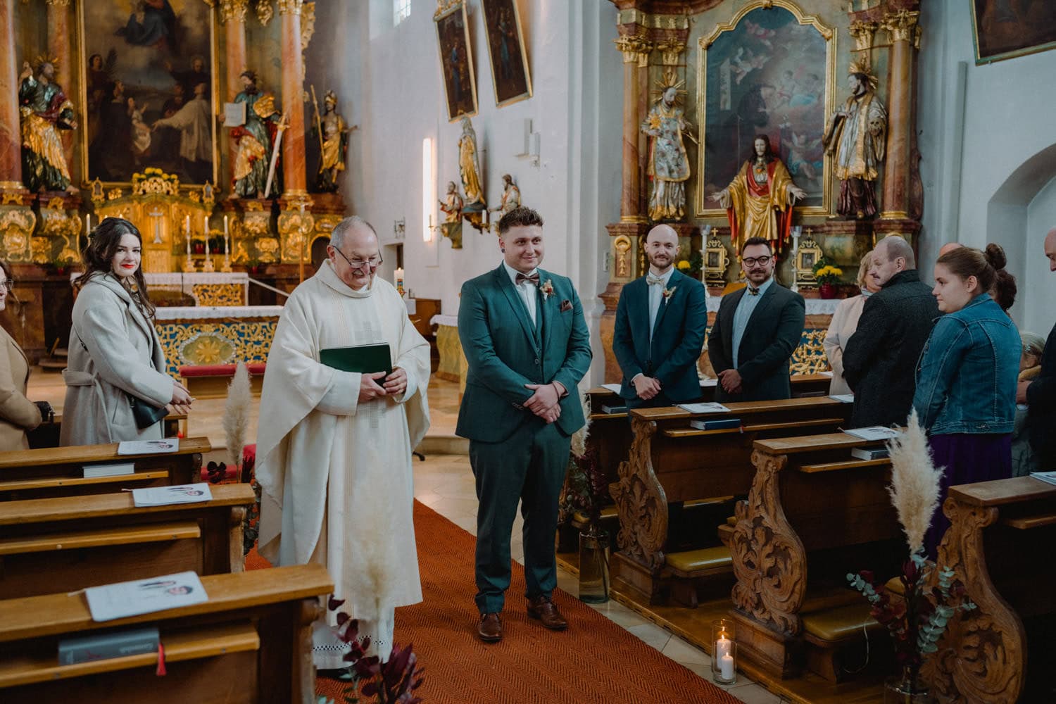 Ein Bräutigam in einem blaugrünen Anzug steht mit einem Priester in der hellen, historischen Hammerschloss Stodl Schmidmühlen-Kirche, umgeben von Gästen, die in reich verzierten Kirchenbänken sitzen. Der Altar ist reich mit religiösen Artefakten und Gemälden geschmückt und schafft eine warme, spirituelle Atmosphäre.