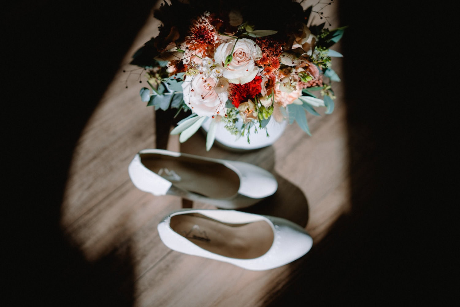 Ein Paar weiße Brautschuhe liegt auf dem Holzboden von Hammerschloss Stodl Schmidmühlen, von sanftem Licht angestrahlt. Darüber hängt ein Strauß gemischter Blumen, rote und weiße Blüten, verflochten mit grünem Blattwerk.
