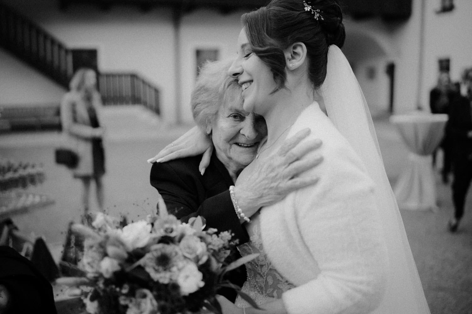 Auf einem zeitlosen Schwarzweißfoto umarmt eine Braut eine ältere Frau herzlich im Hammerschloss Stodl Schmidmühlen. Beide lächeln, während die Braut ihren Blumenstrauß vor einem Hintergrund aus verschwommenen Figuren und einer Außentreppe hält.