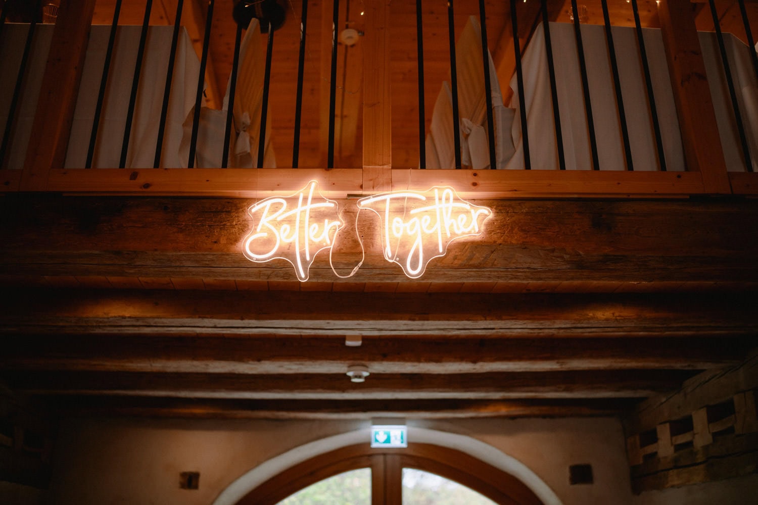Ein Neonschild mit der Aufschrift „Better Together“ hängt an einem rustikalen hölzernen Balkongeländer und wirft einen warmen Schein in den schwach beleuchteten Raum des Hammerschlosses, dessen charmante Holzbalken an den traditionellen Charme von Schmidmühlen erinnern.