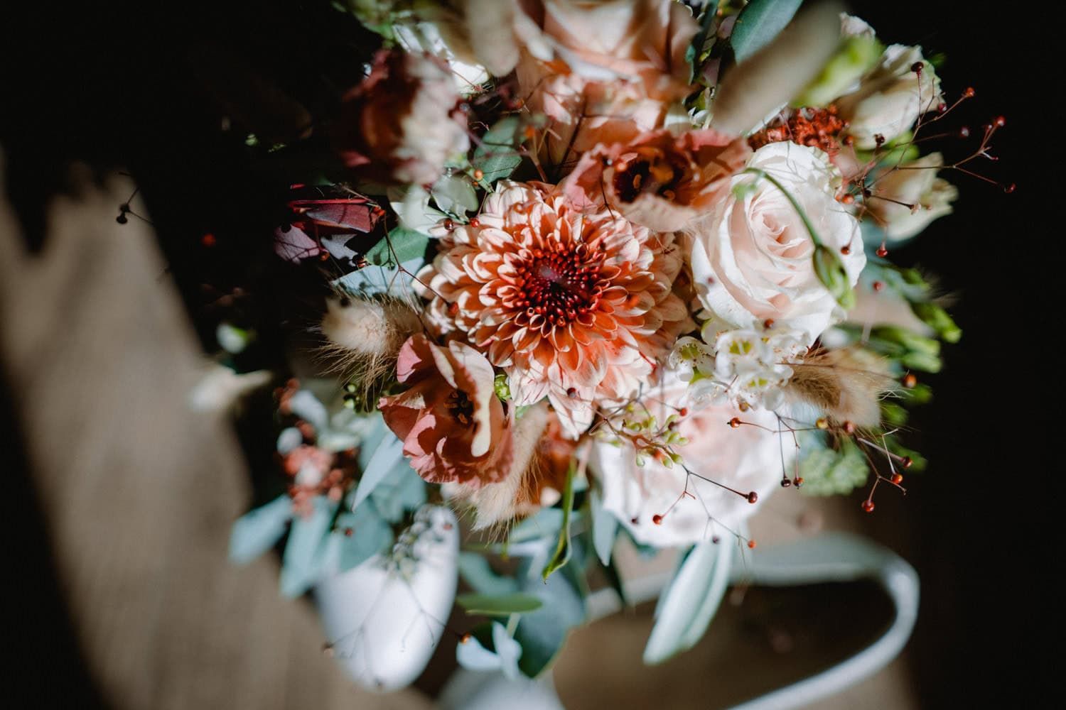 Eine Nahaufnahme eines lebendigen Blumenstraußes mit einer Mischung aus rosa und weißen Rosen von Hammerschloss, einer auffälligen orangefarbenen Dahlie sowie verschiedenen grünen Blättern und kleinen Blüten. Er steht vor einem dunklen Hintergrund, wobei das Sonnenlicht die Blumen hervorhebt und an die zeitlose Eleganz von Schmidmühlen erinnert.
