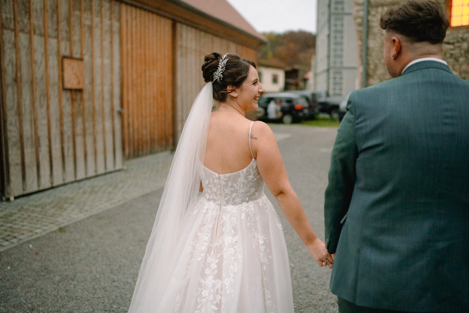 Braut und Bräutigam halten sich an den Händen und schlendern friedlich durch das Gelände von Hammerschloss Stodl Schmidmühlen. Die Braut trägt ein weißes Kleid mit Schleier und der Bräutigam einen grauen Anzug. An diesem ruhigen, bewölkten Tag gehen sie an rustikalen Gebäuden aus Holz und Stein vorbei.
