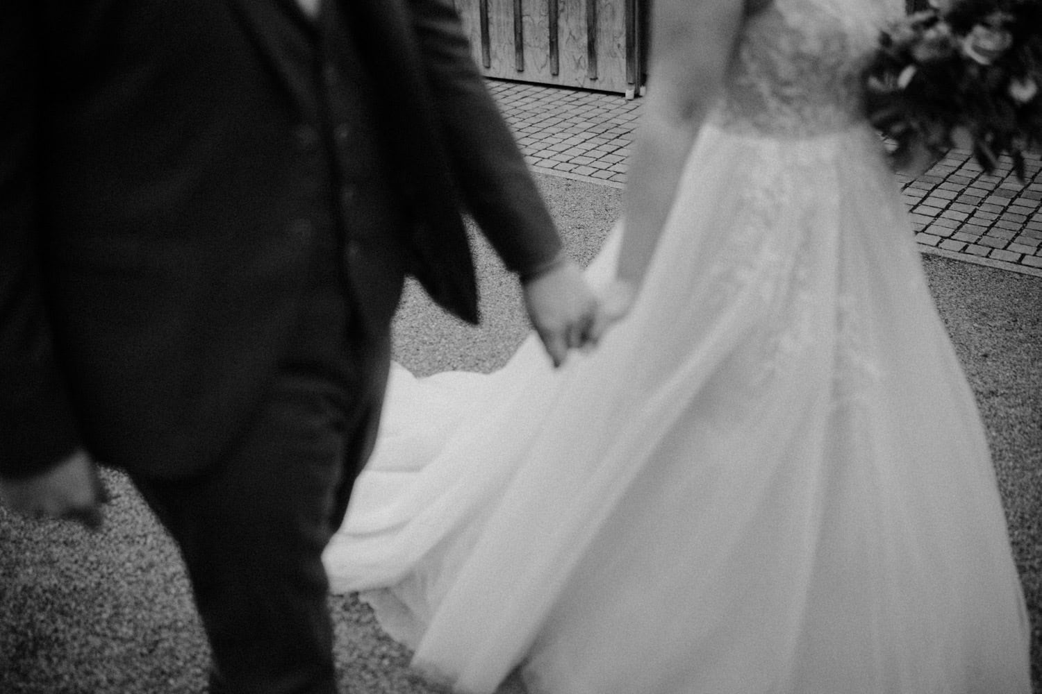 Auf einem Schwarzweißfoto geht ein Paar Hand in Hand davon. Das mit Spitzen verzierte Brautkleid der Frau fließt neben dem Anzug des Mannes. Ihre gefalteten Hände heben sich vor dem verschwommenen Hintergrund aus Gehweg und Holzzaun ab und rufen zeitlose Romantik im Hammerschloss Stodl Schmidmühlen hervor.