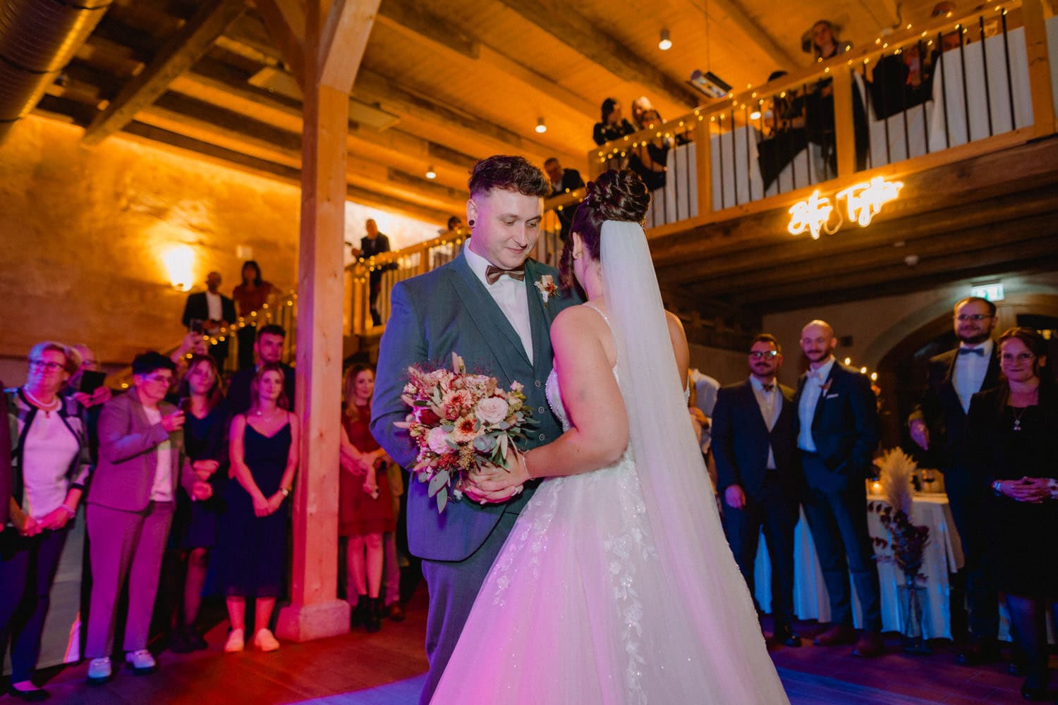 Im charmanten Hammerschloss Stodl Schmidmühlen tanzt ein Paar bei seiner Hochzeitsfeier. Die Braut hält einen Blumenstrauß in der Hand und trägt einen wallenden Schleier, während der Bräutigam in seinem Anzug elegant aussieht. Die Gäste schauen sowohl von der Tanzfläche als auch von einem mit Lichtern geschmückten Balkon im Obergeschoss zu, der von einem leuchtenden Neonschild eingerahmt wird.