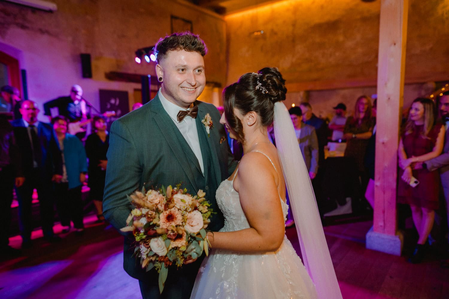 Bei ihrer Hochzeitsfeier im Hammerschloss Stodl tanzen Braut und Bräutigam fröhlich und lächelnd. Der Bräutigam trägt einen dunkelgrünen Anzug, während die Braut elegant ihren Brautstrauß umklammert. Im schwach beleuchteten Hintergrund der bezaubernden Kulisse von Schmidmühlen genießen die Gäste die Feierlichkeiten.