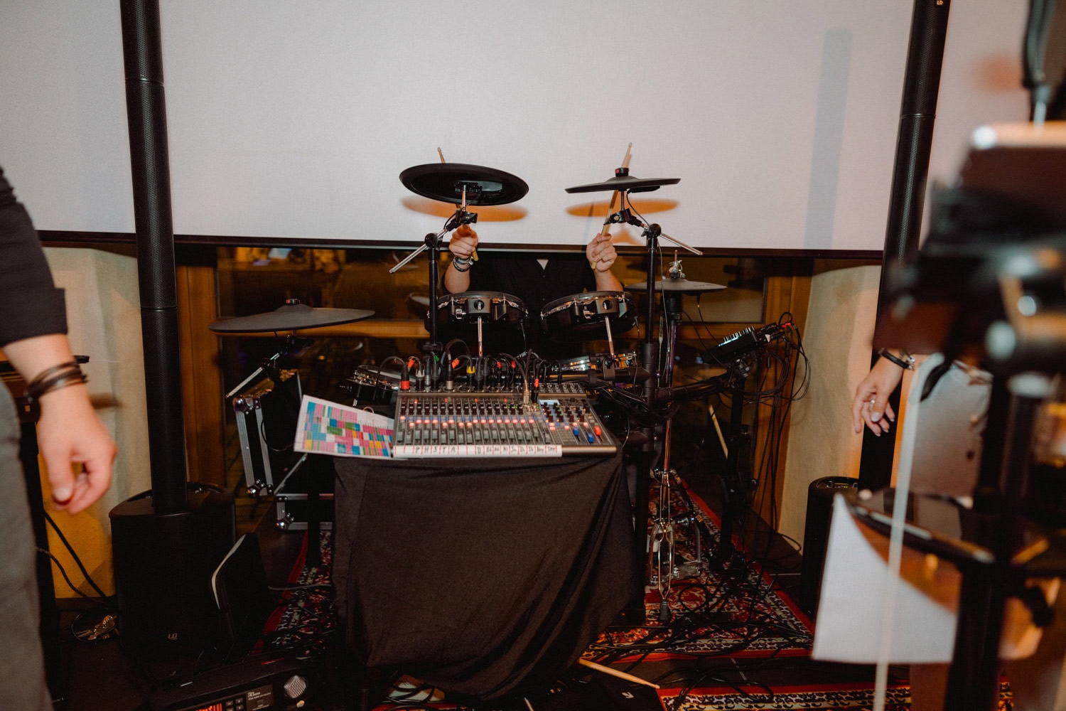 Hinter einem Schlagzeug im Hammerschloss Stodl Schmidmühlen ist ein Schlagzeuger teilweise zu sehen, die Hände mit Trommelstöcken in die Höhe gestreckt. Davor steht ein Mischpult mit bunten Beschriftungen, umgeben von verschiedenen Musikgeräten. Bei zwei weiteren Personen sind die Arme zu sehen, sie sind jedoch größtenteils außerhalb des Bildes.