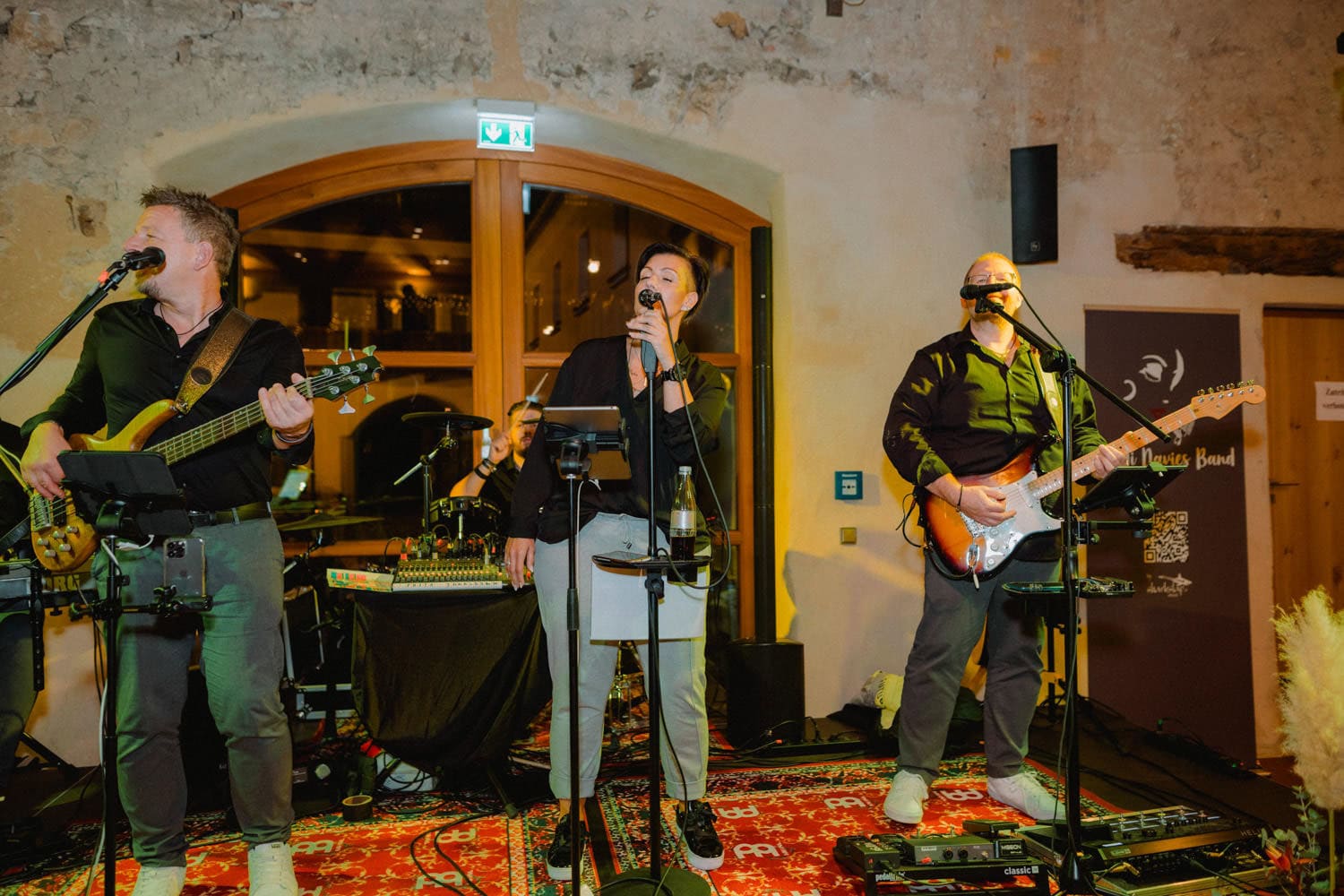Eine Band aus vier Musikern tritt im historischen Hammerschloss Stodl auf. Der Leadsänger steht mit einem Mikrofon in der Mitte der Bühne, flankiert von Gitarristen, während der Schlagzeuger den Beat vor rustikalen Steinmauern und einem großen Fenster in diesem einzigartigen Veranstaltungsort in Schmidmühlen vorgibt.