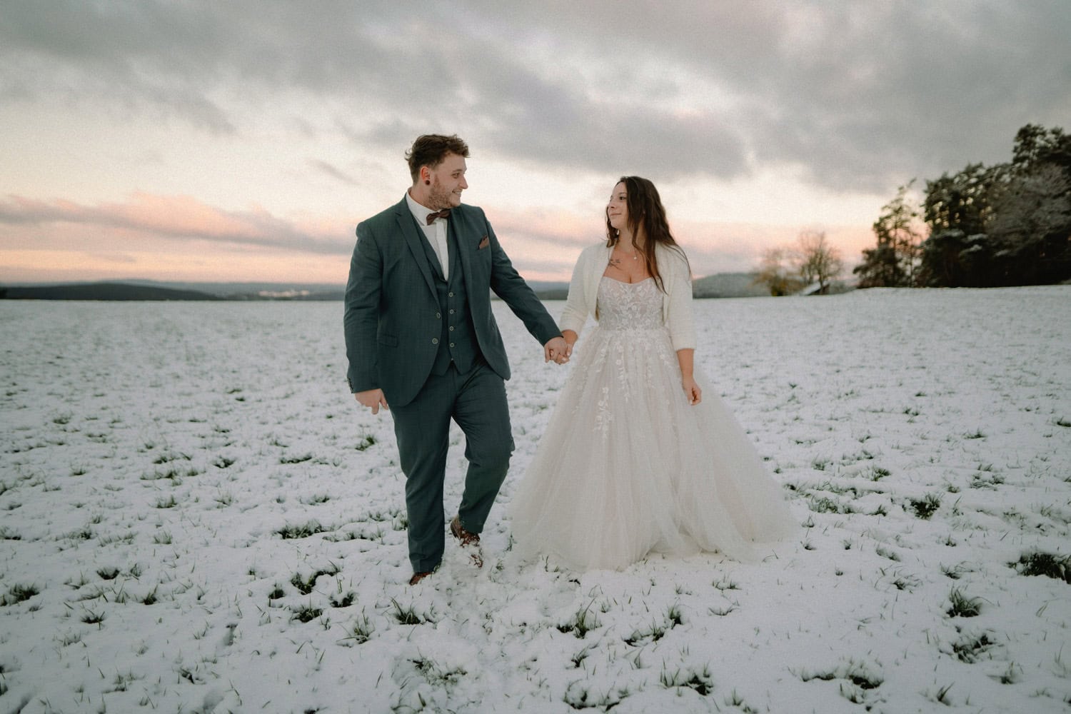 Ein Paar im Hochzeitskleid geht Hand in Hand über ein schneebedecktes Feld in der Nähe von Hammerschloss Stodl Schmidmühlen. Der Bräutigam trägt einen dunklen Anzug und die Braut ein weißes Kleid. Der Himmel ist bewölkt mit einem Hauch von Sonnenuntergang und im Hintergrund sind Bäume zu sehen.