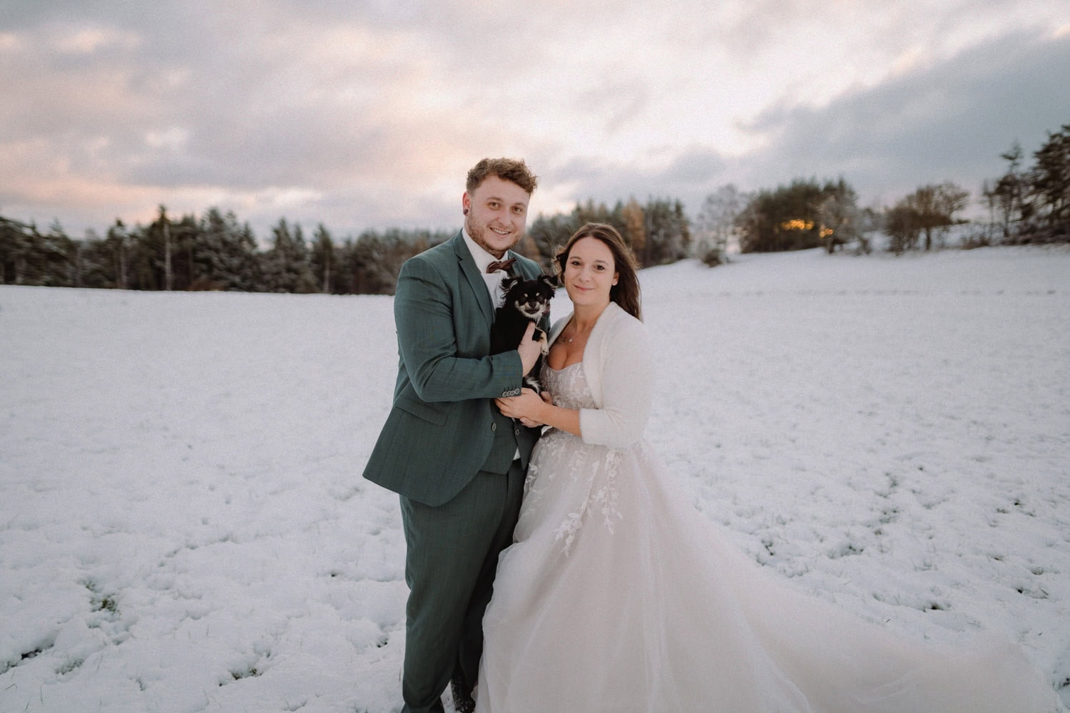Auf dem schneebedeckten Feld von Hammerschloss Stodl steht ein Paar in Hochzeitskleidung zusammen – sie in einem weißen Kleid, er in einem grünen Anzug. Sie lächeln herzlich und halten einen kleinen schwarz-weißen Hund im Arm. Schneebedeckte Bäume und der bewölkte Himmel von Schmidmühlen bieten eine malerische Kulisse.