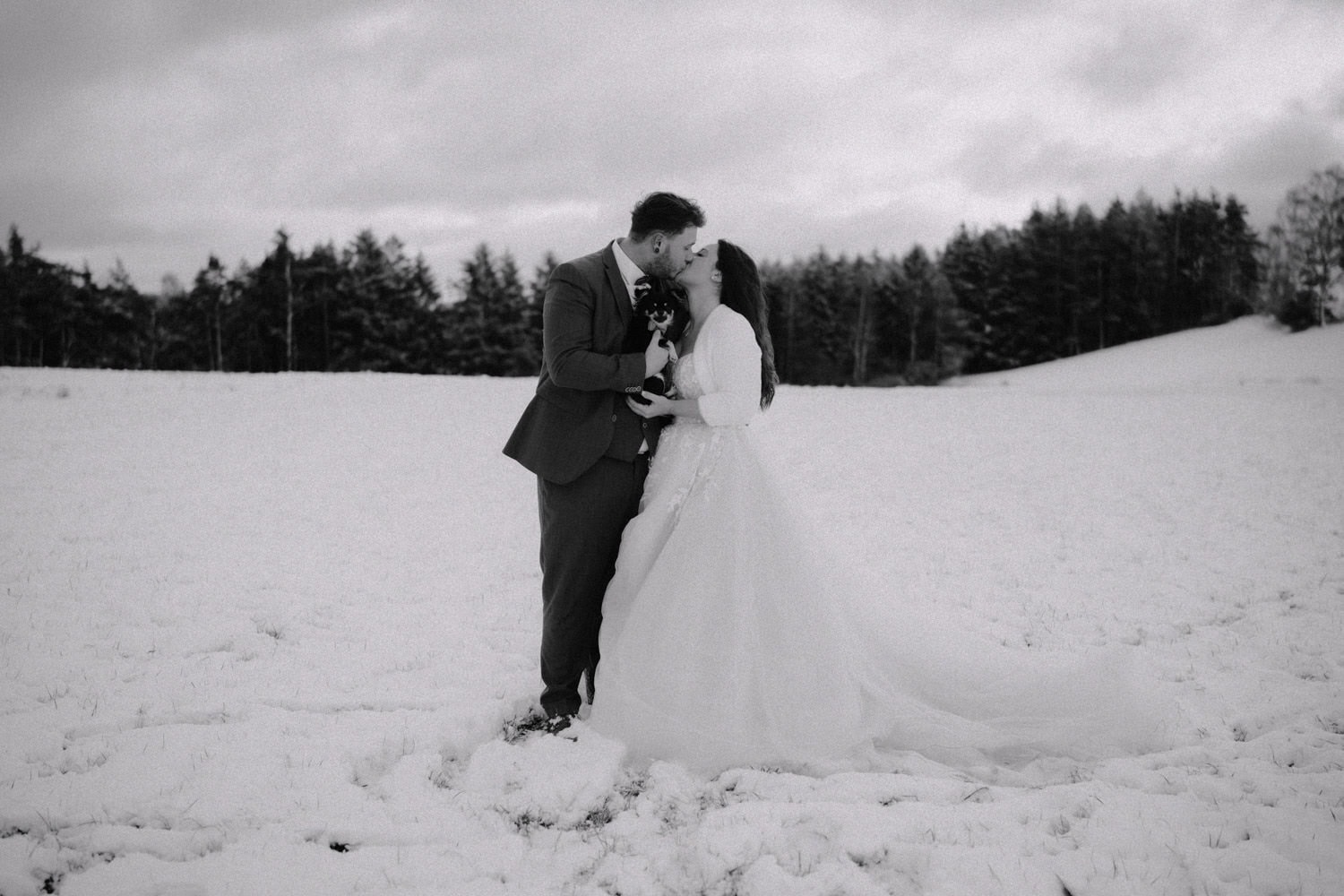 Ein Paar im Hochzeitskleid küsst sich auf einem schneebedeckten Feld beim Hammerschloss Stodl Schmidmühlen und hält einen kleinen Hund. Der Bräutigam trägt einen Anzug und die Braut ein langes Kleid. Bäume säumen den Horizont unter einem bewölkten Himmel.