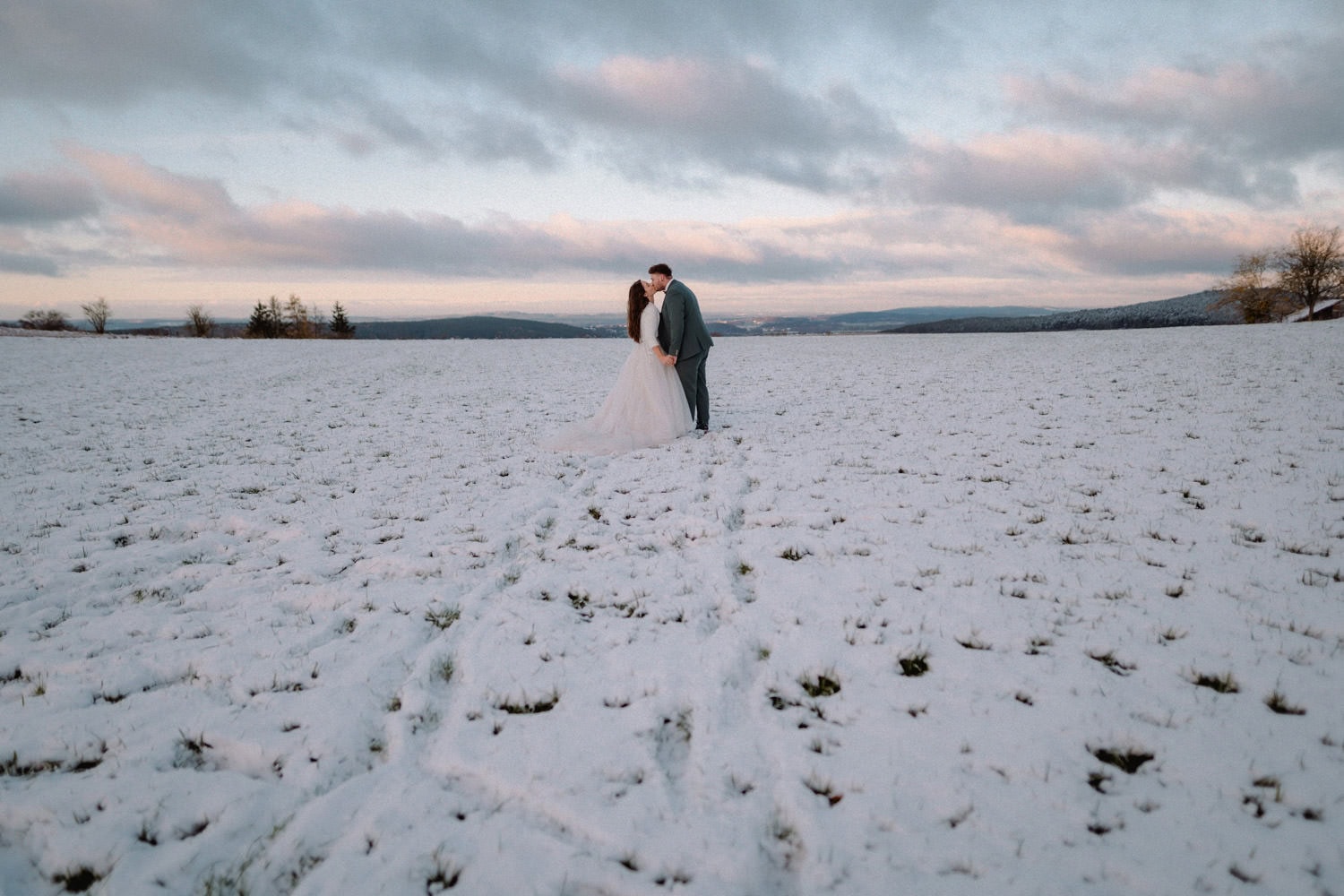 Ein Paar steht und küsst sich inmitten eines schneebedeckten Feldes in der Nähe von Hammerschloss, Schmidmühlen. Der Himmel ist teilweise bewölkt, mit einem Hauch von Blau. Die Landschaft ist ruhig und offen, in der Ferne säumen Bäume den Horizont.