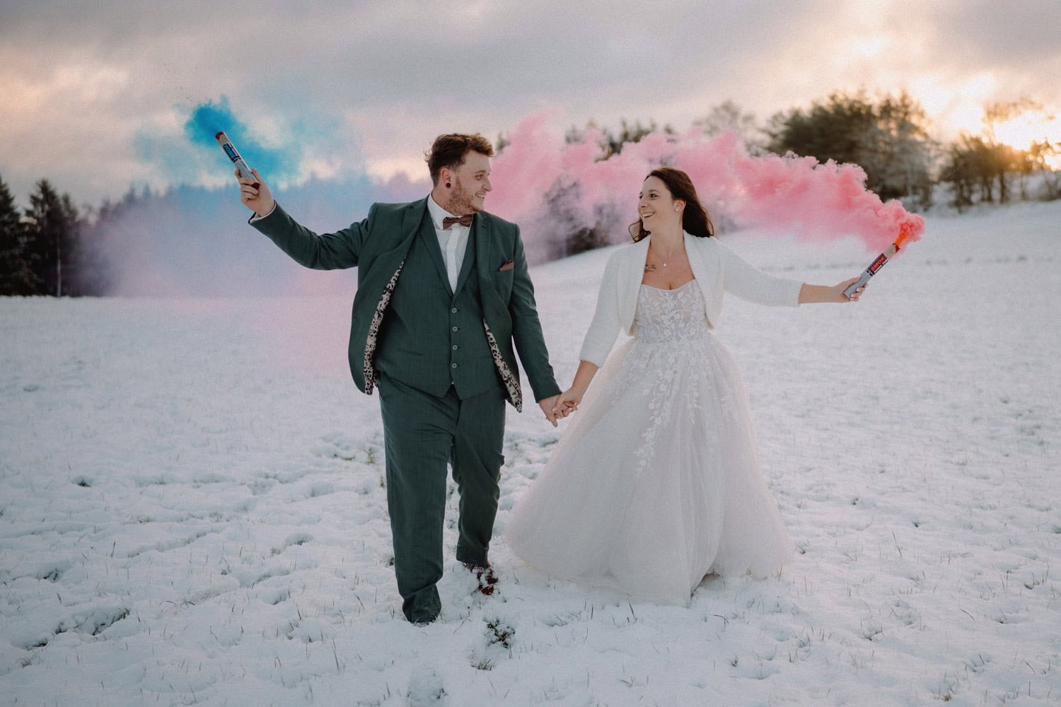 Ein fröhliches Paar im Hochzeitskleid spaziert durch ein verschneites Feld am Hammerschloss und hält Rauchfackeln in der Hand, aus denen rosa und blaue Wolken aufsteigen. Der Bräutigam trägt einen grünen Anzug, während die Braut ein weißes Kleid mit Jacke trägt. Die Sonne geht hinter den verschneiten Bäumen von Schmidmühlen unter.
