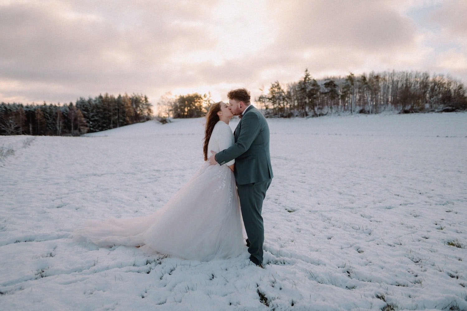 Braut und Bräutigam umarmen sich auf einem schneebedeckten Feld unter einem bewölkten Himmel beim Hammerschloss Stodl Schmidmühlen. Die Braut trägt ein weißes Kleid und der Bräutigam einen Anzug. Hinter ihnen geht die Sonne unter und wirft einen warmen Schein auf die Winterlandschaft mit den Bäumen in der Ferne.