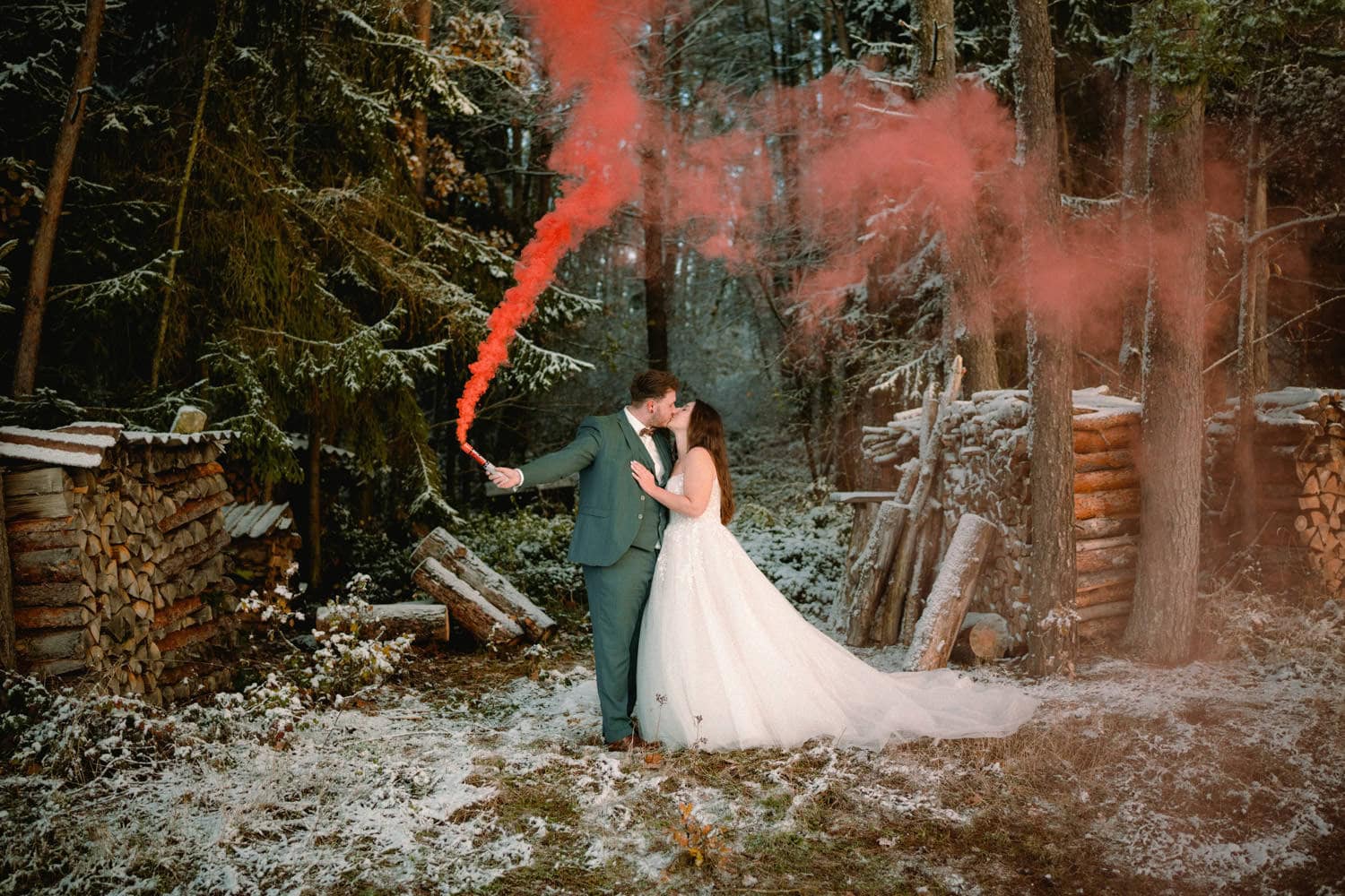 Braut und Bräutigam küssen sich im verschneiten Wald rund um Hammerschloss Stodl Schmidmühlen. Der Bräutigam hält eine rote Rauchfackel in der Hand, die einen leuchtenden Dunst um sie herum erzeugt. Im Hintergrund sind zwischen hohen Bäumen Stapel von Brennholz zu sehen.
