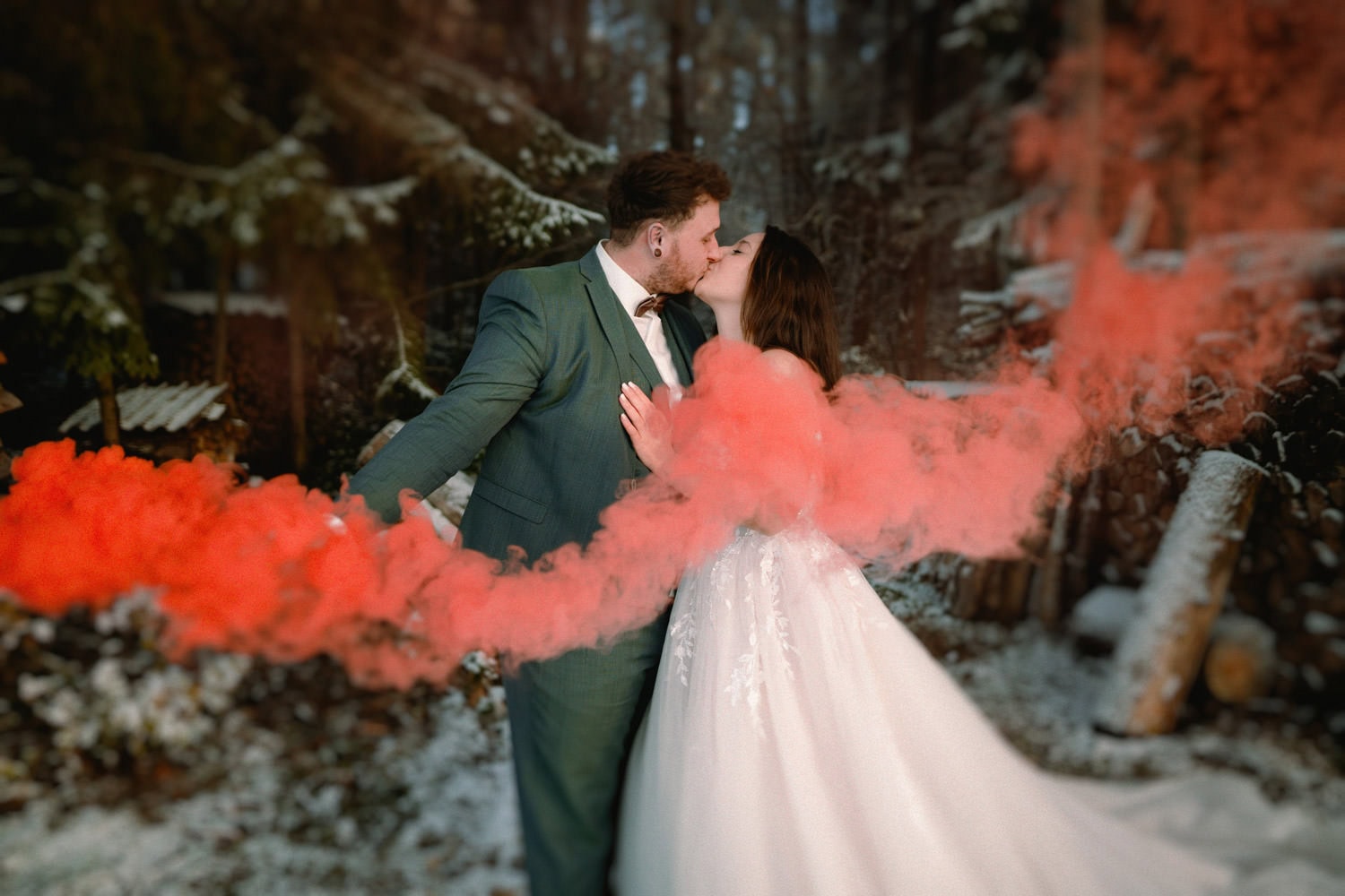 Im verschneiten Wald in der Nähe von Hammerschloss Stodl Schmidmühlen küssen sich Braut und Bräutigam. Der grüne Anzug des Bräutigams bildet einen schönen Kontrast zum weißen Kleid der Braut, während eine rote Rauchwolke um sie herumwirbelt und die dramatische und doch ruhige Winterszene noch verstärkt.