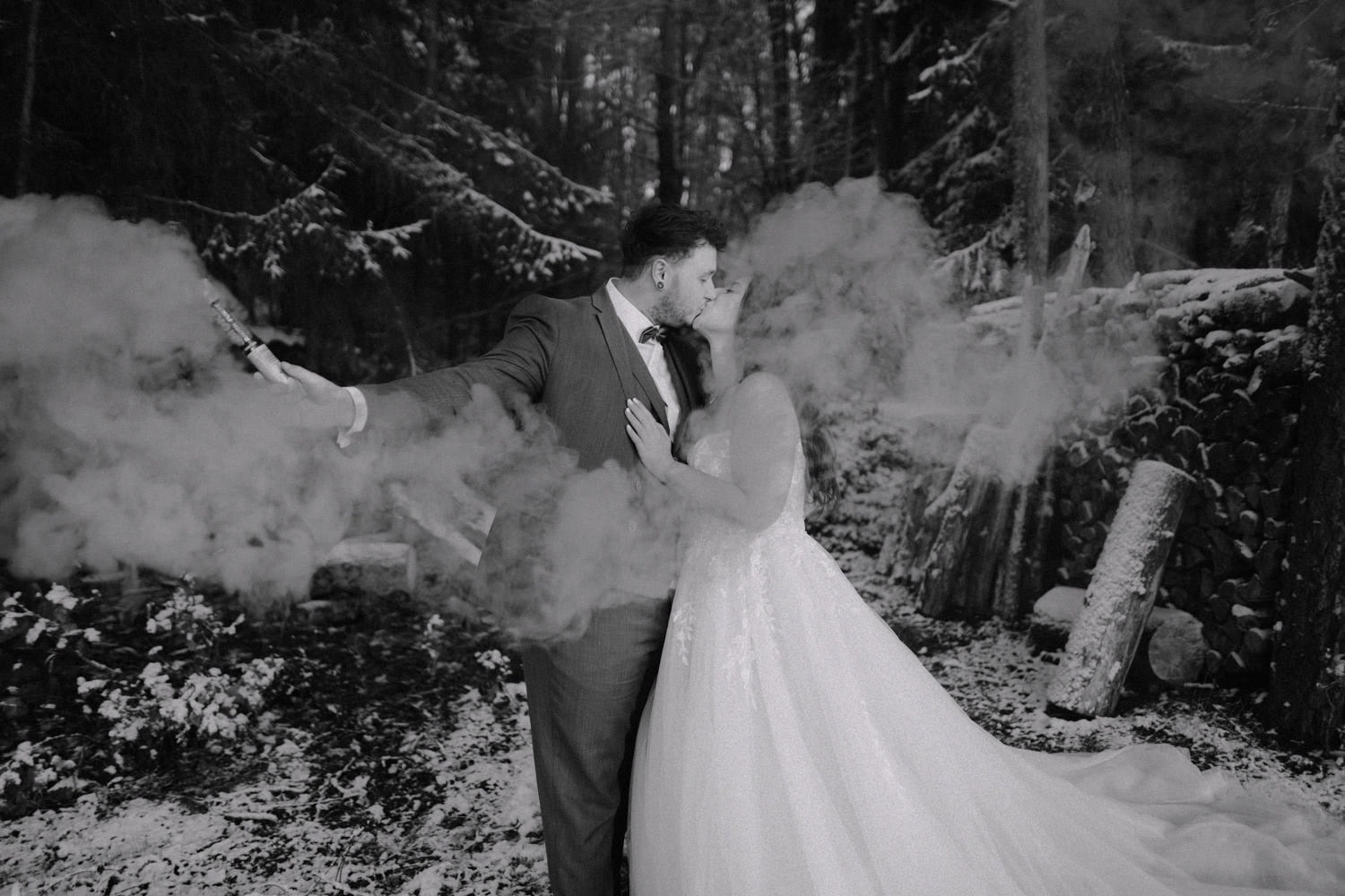 Braut und Bräutigam küssen sich in einem verschneiten Wald in der Nähe von Hammerschloss. Der Bräutigam hält eine Rauchbombe in der Hand, die wirbelnde Wolken um sie herum erzeugt. Die Schwarz-Weiß-Szene zeigt schneebedeckte Bäume und Baumstämme und bietet eine zeitlose Kulisse für ihren intimen Moment.